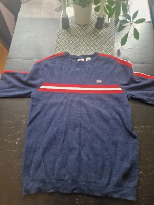 Blå tröja med rödvit rand från Levi's - Mörkblå tröja från Levi's med röda och vita ränder över bröstet och längs ärmarna. Tröjan har rund halsringning och en liten Levi's-logga på bröstet. Perfekt för dig som gillar sportig och retro stil.