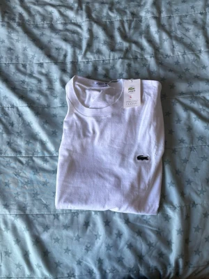 Vit t-shirt från Lacoste  - Klassisk vit t-shirt från Lacoste med rund halsringning och den ikoniska krokodilloggan broderad på bröstet. Tillverkad i mjuk bomull för en skön och stilren look. Perfekt till jeans eller shorts för en clean stil.
