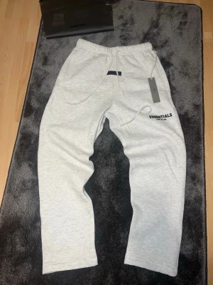 Essentials Fear of God grå mjukisbyxor - Snygga ljusgrå mjukisbyxor från Essentials Fear of God med svart logga på benet. Byxorna har resår i midjan, dragsko och raka ben. Perfekta för en chill och avslappnad stil. Materialet är mjukt och skönt, troligtvis bomullsblandning.