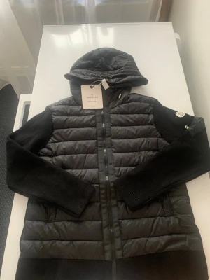 Svart cardiganjacka från Moncler - Svart cardiganjacka från Moncler med quiltad front och huva. Ärmarna är stickade och har en ficka med dragkedja samt Moncler-logga på vänster ärm. Jackan har dragkedja framtill och kombinerar dunmaterial med stickade partier för en modern look. Passar även i XL och M