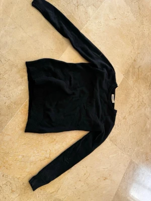 Svart stickad tröja från DBT - En enkel och stilren svart stickad tröja från DBT med rund hals och långa ärmar. Tröjan har ribbstickade muddar vid ärmslut och nederkant, vilket ger en snygg passform. Perfekt för lager-på-lager och passar till många olika outfits.