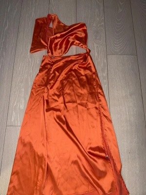 Orange satin one shoulder klänning - Snygg orange långklänning i satin med one shoulder-design och slits på sidan. Klänningen har en asymmetrisk axel och ett glansigt material som ger en lyxig känsla. Perfekt för dig som vill sticka ut med en trendig look.