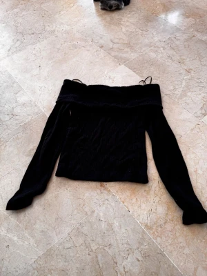 Svart offshoulder ribbad topp - Trendig svart offshoulder topp med långa ärmar och ribbad struktur. Perfekt för dig som vill ha en stilren och enkel look med en twist. Toppen har en bred kant upptill som ger en snygg siluett och sitter skönt på axlarna.