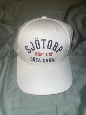 Vit keps med Sjötorp Göta Kanal-tryck - Vit keps med böjd skärm och broderad text 'SJÖTORP', 'N58° E13°' och 'GÖTA KANAL' framtill. Justerbar baktill och ventilerande hål. Snygg och stilren accessoar med marin känsla, perfekt för soliga dagar.