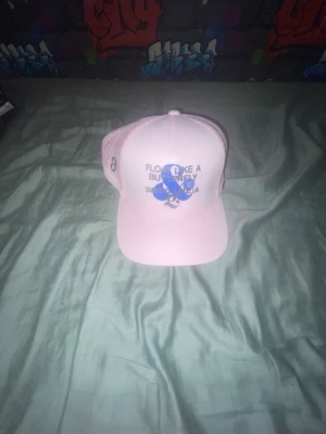 Rosa keps från G/FORE med tryck - Säljer en ljusrosa keps från G/FORE med blått tryck framtill där det står 'Float like a butterfly, swing like a G'. Kepsen har mesh på sidorna och justerbar snapback baktill. Perfekt för dig som gillar sportig och trendig stil.