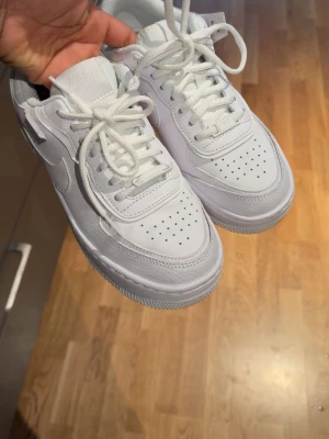 Nike Air Force 1 Shadow vita sneakers - Nike Air Force 1 Shadow i helvitt läder med klassisk chunky sula och dubbla detaljer på swoosh och häl. Skorna har perforerad tå, vita skosnören och en clean look som passar till allt. Perfekta för dig som gillar streetstyle och vill sticka ut lite extra.
