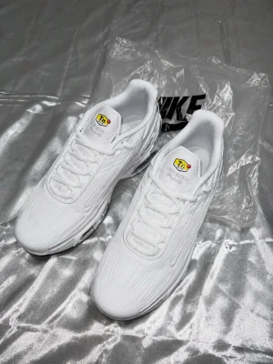 Nike Air Max Plus Vita Sneakers - Nike Air Max Plus sneakers i helvitt med tydliga TN-loggor på plösen och sidan. Skorna har en sportig siluett, synliga Air Max-enheter i sulan och detaljerad textur på ovandelen. Perfekta för dig som gillar en clean och modern look.