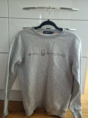 Grå sweatshirt från Sail Racing - Snygg grå sweatshirt från Sail Racing med tryckt logga på bröstet. Tröjan har rund halsringning, långa ärmar och är gjord i mjukt bomullsmaterial. Perfekt för en avslappnad och sportig stil.