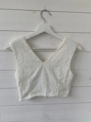 Vit spetsig croppad linnetopp - Snygg vit croppad linnetopp med v-ringning och spetsdetaljer. Toppen är ärmlös och har en feminin, elegant look som passar perfekt till både jeans och kjol. Perfekt för dig som gillar en stilren och fräsch vibe.