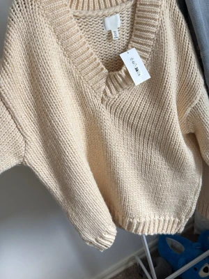 Beige stickad tröja från H&M - Säljer en chunky beige stickad tröja från H&M med djup v-ringning och bred ribbad kant vid halsen. Tröjan har avslappnad passform, långa ärmar och ribbade muddar. Perfekt för lager på lager och riktigt cozy vibes.