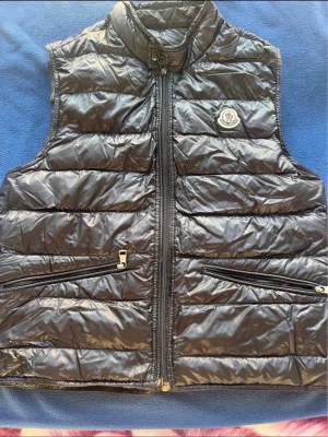  Moncler väst  - Pris diskuteras vid snabb affär. Snygg svart dunväst från Moncler med glansig finish och klassisk logotyp på bröstet. Västen har hög krage, två dragkedjeförsedda fickor framtill och quiltad design. Perfekt för lager-på-lager och streetstyle.