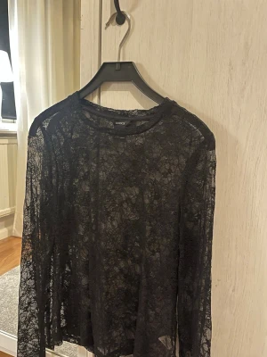 Svart spetsblus från Lindex - Snygg svart långärmad blus från Lindex i transparent spetsmaterial. Perfekt att styla lager på lager eller med ett linne under. Blusen har rund halsringning och ett elegant, feminint uttryck.