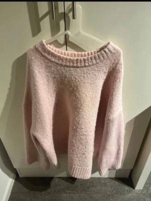 Ljusrosa fluffig stickad tröja - Supermjuk och oversized stickad tröja i ljusrosa med bred ribbad kant vid hals, ärmslut och nederkant. Perfekt för dig som gillar en avslappnad och cozy stil. Tröjan har en enkel design utan synligt märke och är gjord i ett fluffigt material.
