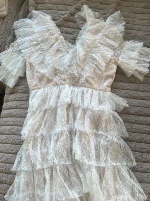 Vit By Malina klänning  - Superfin vit klänning med flera lager volanger och spetsdetaljer. Klänningen har v-ringning och korta ärmar med volang. Perfekt för dig som vill sticka ut med en romantisk och luftig look. Materialet är spets och klänningen har en ljus ton.