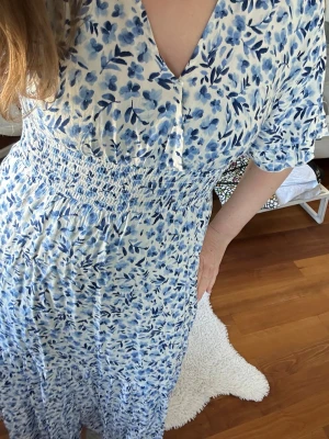 Blommig blåvit långklänning - Snygg långklänning med blått blommönster.Klänningen har v-ringning, kort ärm och smock i midjan för en bekväm passform. Perfekt för varma dagar och somriga tillfällen.