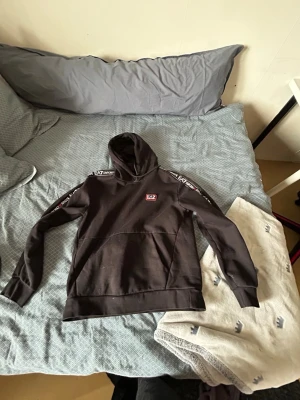 Svart hoodie från EA7 Emporio Armani - Svart hoodie från EA7 Emporio Armani med logga på bröstet och tryck längs ärmarna. Klassisk känguruficka framtill och huva. Perfekt för en avslappnad och sportig stil.