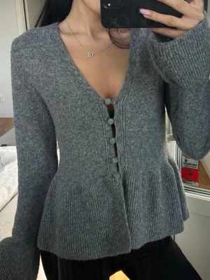Grå peplum kofta med knappar - Mysig grå kofta med peplumskärning och stora tygklädda knappar framtill. Tröjan har v-ringning, långa ärmar och ribbstickade detaljer vid ärmslut och nederkant. Perfekt för dig som gillar en feminin och trendig look.