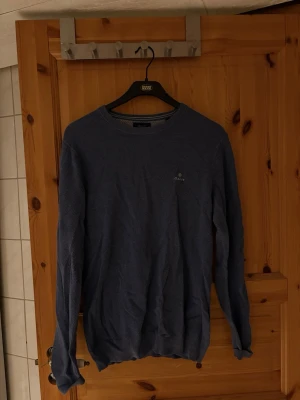 Blå stickad tröja från Gant - Snygg blå stickad tröja från Gant med rund halsringning och diskret logga broderad på bröstet. Tröjan har en klassisk passform och ribbade muddar vid ärmslut och nederkant. Perfekt för en stilren look.