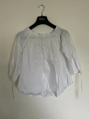 Vit offshoulder blus med resår - Snygg vit offshoulder blus från LOGG med smockad resår upptill och trekvartsärmar med knytband. Blusen är luftig och har en avslappnad passform, perfekt till jeans eller kjol. Tillverkad i ett lätt och mjukt tyg.