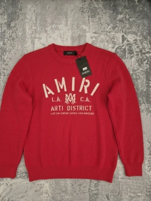 Amiri Röd stickad tröja - Snygg röd stickad tröja från Amiri med vit text och logga framtill. Rund halsringning och ribbade muddar vid ärmslut och nederkant. Perfekt för dig som gillar streetwear och vill ha något trendigt och bekvämt. Passar medium och small