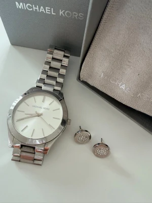 Silverfärgad klocka från Michael Kors+Örhängen Michael Kors - Stilren klocka från Michael Kors i silverfärgat metallarmband och rund urtavla med minimalistisk design. Klockan har en klassisk look med diskreta index och logga på urtavlan. Perfekt accessoar för en trendig och modern stil.