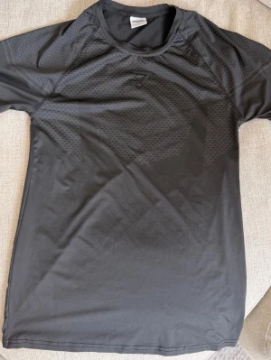 Svart träningströja från Gymshark - Svart kortärmad träningströja från Gymshark med diskret mönster över bröstet och axlarna. Tillverkad i ett lätt och stretchigt funktionsmaterial som andas, perfekt för intensiva träningspass. Snygg och sportig design med rund hals.