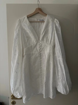Vit kortklänning från Adoore - Superfin vit kortklänning från Adoore med broderade blommor, v-ringning och långa puffärmar. Klänningen har knappar framtill och är tillverkad i ett lätt material som känns luftigt och fräscht. Perfekt för dig som gillar detaljer och stilren design.