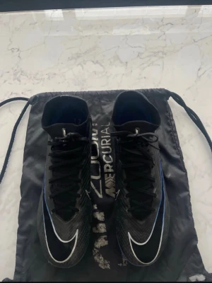 Nike Mercurial Air Zoom fotbollsskor - Säljer ett par Nike Mercurial Air Zoom fotbollsskor i svart med blå och vita detaljer. Skorna har en strömlinjeformad design med högt skaft och snörning. Ovandelen är i syntetmaterial och sulan har fasta dobbar för spel på gräs. Väskan följer med