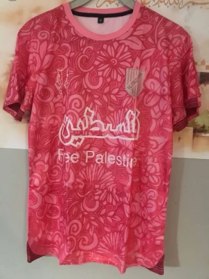 Rosa fotbollströja Free Palestine/ xl  - Säljer en rosa fotbollströja med blommigt mönster och texten 'Free Palestine' på bröstet. Tröjan har korta ärmar, rund halsringning och klubbmärke på vänster sida. Perfekt för dig som vill sticka ut på planen.