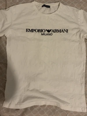 Vit Emporio Armani t-shirt - Vit t-shirt från Emporio Armani med svart logotyp och texten 'Milano' tryckt på bröstet. Klassisk rund halsringning och korta ärmar. Perfekt för en stilren och trendig look.