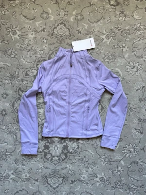 Ljuslila träningsjacka från Lululemon - Snygg ljuslila träningsjacka från Lululemon med figurnära passform, hel dragkedja framtill och två fickor med dragkedja. Jackan har hög krage och långa ärmar, perfekt för träning eller powerwalks. Tillverkad i ett stretchigt funktionsmaterial.