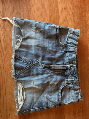 Blå jeanskjol med slitna detaljer - Säljer en kort blå jeanskjol med råa, fransiga kanter och slitna detaljer. Kjolen har klassiska fickor fram och bak samt bälteshällor. Perfekt för dig som gillar en avslappnad och trendig stil.