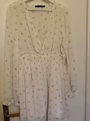 Vit blommig kortklänning  - Söt vit kortklänning  med små blommor i pastellfärger. Klänningen har v-ringning, långärmade ärmar med resår vid handlederna och smock i midjan för en bekväm passform. Perfekt för vår och sommar!