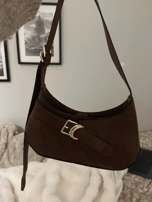 H&M Brun axelremsväska i mocka - Snygg brun handväska i mocka från H&M med justerbar axelrem och guldfärgat spänne framtill. Väskan har en mjuk, rundad form och minimalistisk design som passar till många olika stilar. Perfekt accessoar för dig som gillar klassiska detaljer.