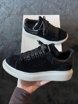 Svarta sneakers från Alexander McQueen - Stilrena svarta sneakers från Alexander McQueen med breda svarta skosnören, mockaliknande ovandel och vit chunky sula. Märkeslogga broderad på plösen och hälparti i svart läder. Perfekta för dig som gillar exklusiv streetstyle.