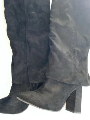 Svarta höga stövlar med klack - Säljer ett par svarta höga stövlar i om mockaimitation med bred klack och spetsig tå. Stövlarna har dragkedja på insidan och en stilren look som passar till många outfits. Perfekta för dig som vill ha en edgy och trendig stil.