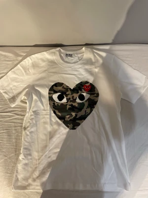 Vit Comme des Garçons PLAY t-shirt - Vit t-shirt från Comme des Garçons PLAY med stort hjärta i camouflage-mönster och ögon på bröstet. Liten röd hjärtdetalj med ögon ovanpå. Klassisk passform och rund hals. Perfekt statement-plagg för dig som gillar streetwear.