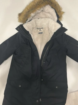 Svart Vinterjacka från McKinley - Svart Vinterjacka från McKinley med mysigt vitt teddyfoder och huva med fuskpäls. Jackan har dragkedja, tryckknappar och stora fickor framtill. Perfekt för kalla dagar med sin långa modell och varma material.