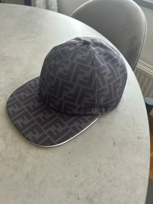 Svart keps med Fendi-logga - Säljer en svart keps från Fendi med det klassiska FF-monogrammet över hela kepsen. Kepsen har rak skärm och är tillverkad i ett slitstarkt tygmaterial. Perfekt för dig som vill ha en stilren och trendig accessoar med lyxig känsla.