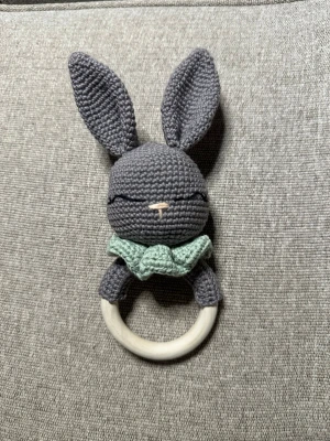 Virkad kanin skallra med trähandtag - Söt handgjord skallra i form av en kanin med grått virkat huvud, långa öron och mintgrön krage. Skallran har ett runt handtag i trä som är lätt att greppa. Perfekt som leksak till små barn eller som present.