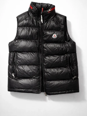 Svart dunväst från Moncler - Snygg svart dunväst från Moncler med inbyggd luva. Västen har två dragkedjefickor och Moncler-logga på bröstet. Insidan har en unik serietecknad tvättetikett. Perfekt för lager-på-lager och streetstyle.