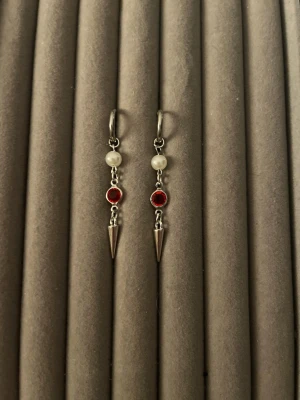 Silvriga örhängen med pärlor och röd sten - Säljer ett par snygga silvriga örhängen med vita pärlor, röda stenar och spetsiga hängen. Örhängena har en cool och modern design som sticker ut. Perfekta för dig som vill ha något unikt och trendigt i smyckeskrinet.
