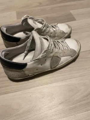 Vita låga sneakers från Philippe Model - Snygga vita sneakers från Philippe Model med beige mockadetaljer och svart häl. Skorna har klassisk låg profil, vita snören och en ikonisk sköld på sidan. Materialmix av läder och mocka ger en schysst kontrast och streetkänsla.