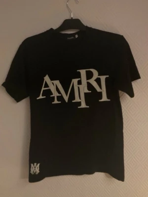 Svart AMIRI t-shirt med stor logga - Snygg svart t-shirt från AMIRI med stor vit logga framtill och en mindre logga nere vid fållen. Klassisk rund hals och korta ärmar. Perfekt för dig som gillar streetwear och vill sticka ut med en bold design.