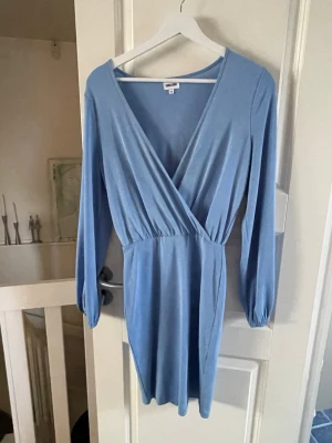 Ljusblå omlottklänning med v-ringning - Snygg ljusblå kortklänning med omlott och v-ringad front. Klänningen har långa ärmar och resår i midjan för en smickrande passform. 