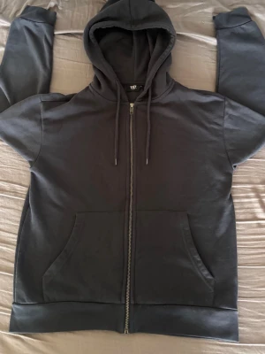 Mörkgrå zip hoodie med huva - En enkel och stilren mörkgrå hoodie med dragkedja framtill och två stora fickor. Hoodien har justerbar huva med snören och ribbade muddar vid ärmslut och nederkant. Perfekt för en avslappnad look och passar till det mesta. Märke: lager 157. Storlek: S(känns som xs)