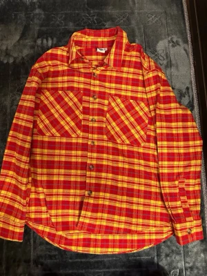 Saint Vanity skjorta - Riktigt snygg röd och gul rutig skjorta från Saint Vanity med stora bröstfickor och coolt tryck på ryggen. Skjortan har klassisk krage, knappar framtill och lång ärm. Perfekt för dig som vill sticka ut med en unik streetwear-look.