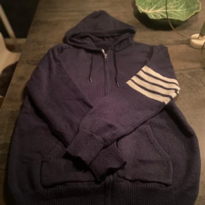 Marinblå stickad hoodie - Mysig marinblå stickad hoodie med huva och dragkedja. Ena ärmen har vita ränder och på ryggen finns ett stort segelbåtsmotiv i vitt och rött. Perfekt för dig som gillar en chill och nautisk stil.