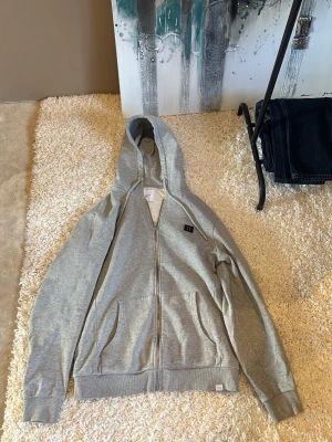 Grå zip hoodie från Les Deux - Snygg grå hoodie från Les Deux med dragkedja framtill och två fickor. Klassisk huva med dragsko och mjukt, fleecefodrat innerfoder för extra komfort. Perfekt för en avslappnad streetwear-look.