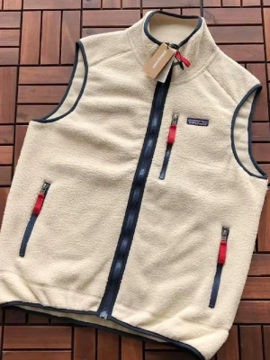 Beige fleeceväst från Patagonia - Snygg beige fleeceväst från Patagonia med marinblå detaljer och dragkedjor. Västen har tre fickor med dragkedja, varav två framtill och en på bröstet. Mjukt och mysigt material, perfekt för lager-på-lager och outdoor-vibe.